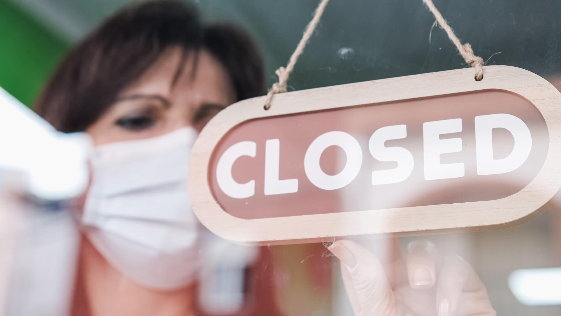 Woman wearing a face mask while turning a sign from open to closed through window. Office losed